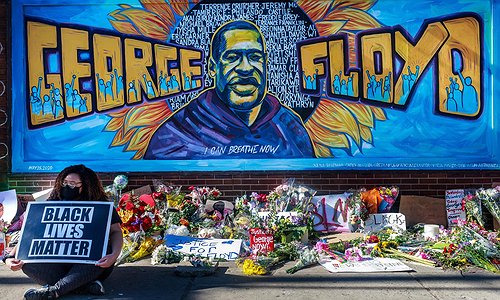 A colorful mural of George Floyd who was unjustly killed by police in Minneapolis. His face is in the middle with the words George Floyd in large, orange block lettering. the text "Say our names" and "I can breathe now" is written above and below George's head with the names of fellow Black people who have been killed by police written behind him.