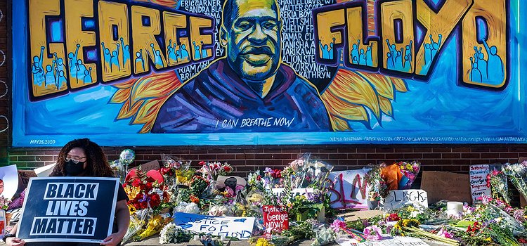 A colorful mural of George Floyd who was unjustly killed by police in Minneapolis. His face is in the middle with the words George Floyd in large, orange block lettering. the text "Say our names" and "I can breathe now" is written above and below George's head with the names of fellow Black people who have been killed by police written behind him.