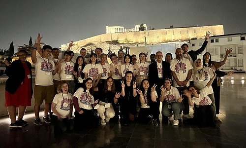 A picture of some of the organizing committee and student volunteers at the Acropolis Museum with the lit up Parthenon in the background