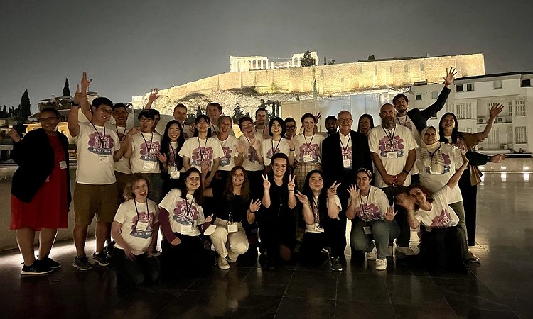 A picture of some of the organizing committee and student volunteers at the Acropolis Museum with the lit up Parthenon in the background