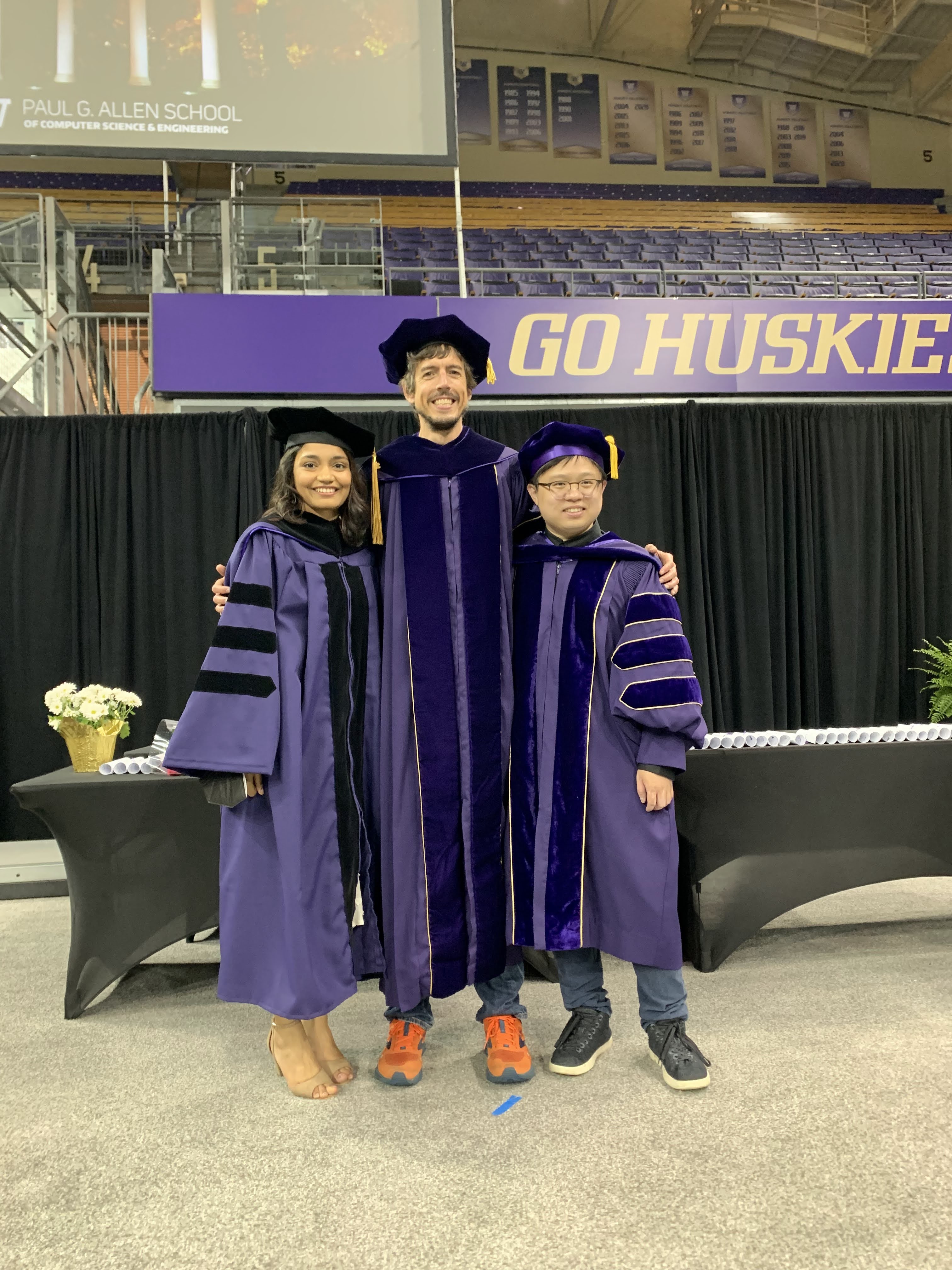 Manaswi, Jon, and Liang standing with their graduation gowns in front of the "Go Huskies! sign